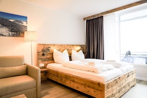 Un dormitorio con cama de madera, una silla, una mesita de noche, una lámpara y una ventana con cortinas.