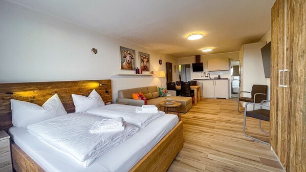 A hotel room with a large bed, a sofa, a small table, and a kitchenette in the background.