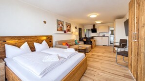 A hotel room with a wooden headboard, a bed with white linens, a sofa, a small table with a lamp, and a kitchenette in the background.