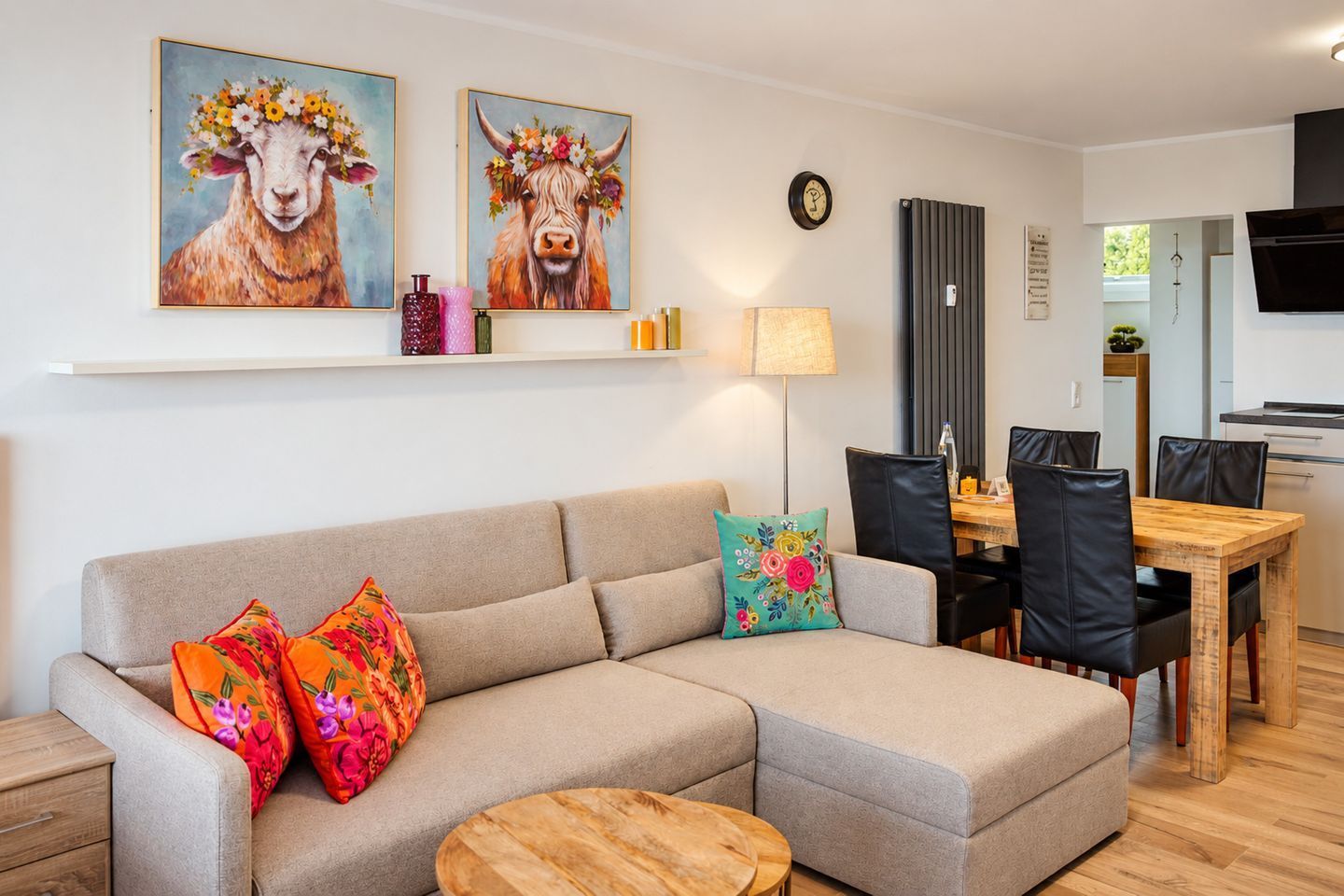 Una sala de estar con un sofá, dos almohadones coloridos, una mesa de centro de madera, un área de comedor con una mesa y sillas de madera, y dos cuadros de animales con coronas de flores.
