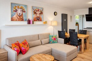 Una sala de estar con un sofá, dos almohadones coloridos, una mesa de centro de madera, un área de comedor con una mesa y sillas de madera, y dos cuadros de animales con coronas de flores.