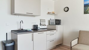 Eine kleine Küchenzeile mit Mikrowelle, Wasserkocher und Geschirr auf der Arbeitsplatte, einem Mülleimer und einem Stuhl in der Ecke.