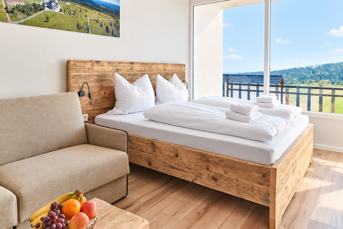 Ein Schlafzimmer mit einem hölzernen Bett, einem Schlafsofa und einem großen Fenster mit Blick auf einen Balkon und viel Grün.