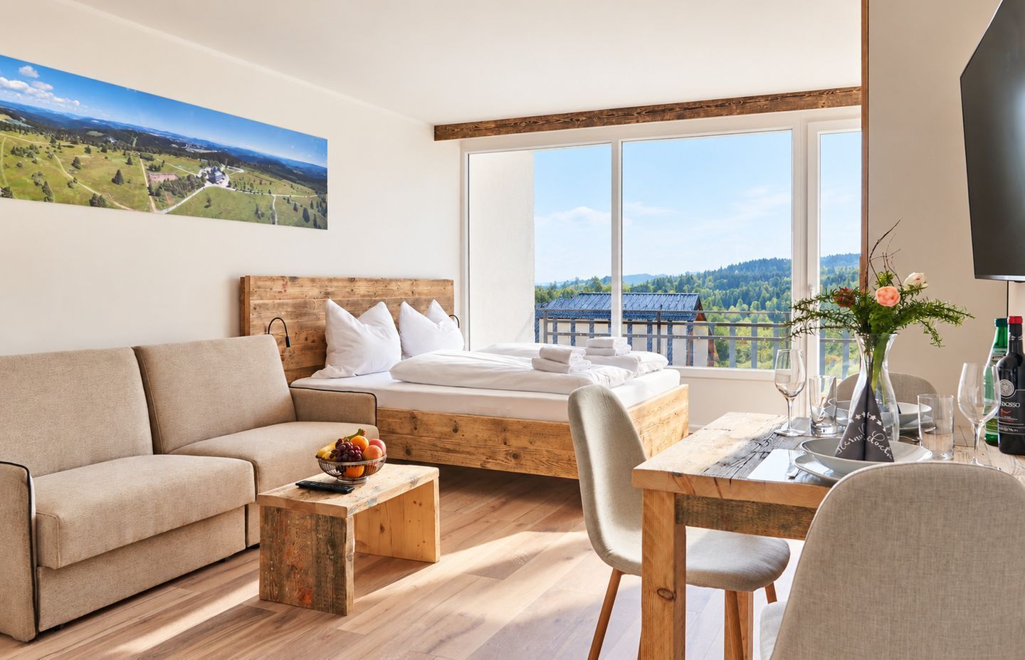 A modern hotel room with a bed, sofa, dining table, and a large window offering a scenic view.