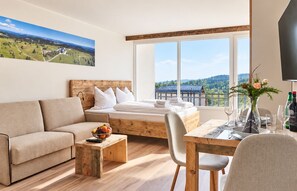 A modern hotel room with a bed, sofa, dining table, and a large window offering a scenic view.