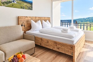 A bedroom with a wooden bed, a sofa, and a large window with a view of a balcony and greenery. - Smart Resorts - House Brilliant - Studio apartment 609 with balcony, max. 4 persons (Winterberg)