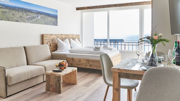 Ein modernes Hotelzimmer mit Bett, einer Couch, einem Esstisch und Blick auf die Landschaft.