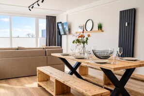 A modern living room with a wooden dining table, bench seating, a sofa, and a flat-screen TV mounted on the wall. - Smart Resorts - Haus Brilliant - Ferienwohnung 602 mit 3 Schlafzimmer und Balkon, Max. 8 Personen - (Winterberg)