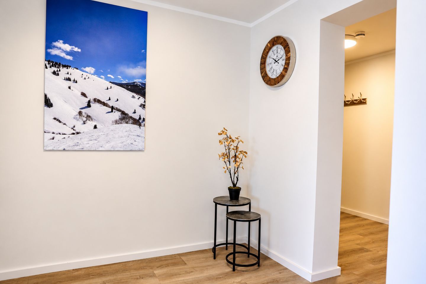 A hallway with a wooden floor, a wall-mounted clock, a framed landscape photo, and a small table with a potted plant.