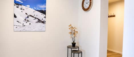 Un pasillo con piso de madera, un reloj en la pared, una fotografía enmarcadas de un paisaje y una pequeña mesa con una planta en maceta.