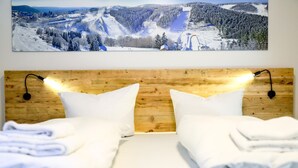 A hotel room with a wooden headboard, two white pillows, a neatly made bed with white linens, and a large landscape painting of a snowy mountain scene above the bed.