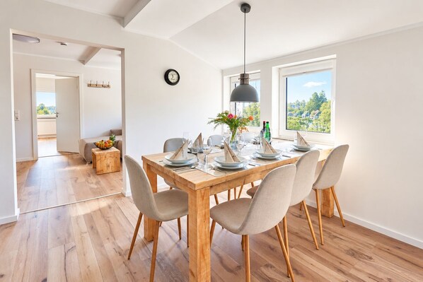 A dining area with a wooden table set for six, a modern pendant light, and a view of greenery outside.