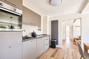 A modern kitchen with a built-in oven, a coffee machine, and a trash bin. There is a dining area with a wooden table and chairs visible in the background.