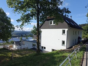 Une maison blanche à deux étages, avec un toit en pente, entourée d’une clôture en bois et d’un arbre.