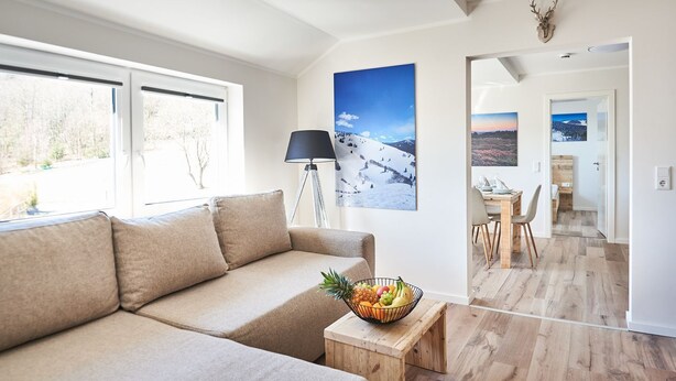 Ein modernes Wohnzimmer mit einem beigefarbenen Sofa, einem Couchtisch mit Obstschale, einer Stehlampe und einem großen Fenster.