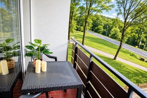 A balcony with a table, chairs, potted plants, and a view of a road and greenery.