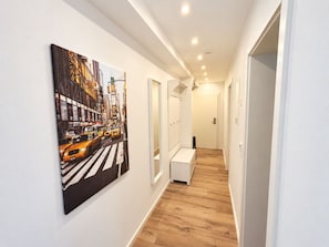 Un pasillo con un cuadro de una calle urbana, un banco blanco y una puerta al final.
