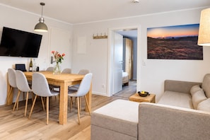 A modern living room with a wooden dining table, grey upholstered chairs, a sofa, a flat-screen TV, and a painting of a sunset landscape. - Smart Resorts - Haus Saphir - Ferienwohnung 504 mit 2 Schlafzimmern, Max. 7 Personen - Smart Resorts (Winterberg)