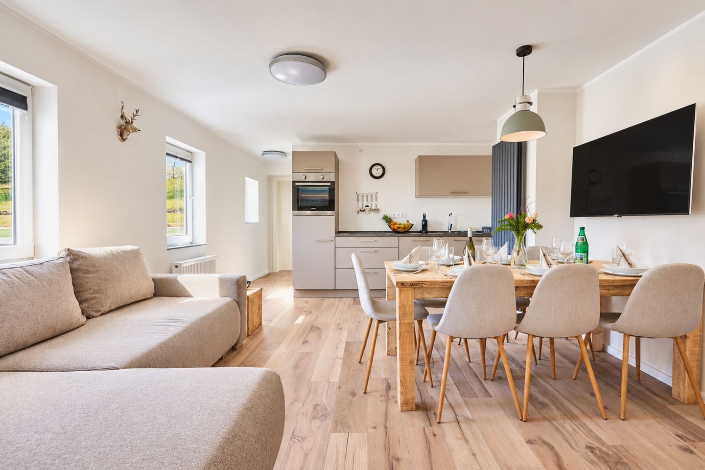 A modern living room with a sofa, dining table with chairs, a kitchen area with a TV, and a clock on the wall.