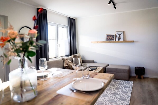 A dining area with a wooden table set for four, a sofa, and a wall-mounted shelf with framed pictures.