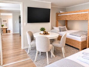 Une chambre moderne avec un lit superposé, une table de salle à manger ronde entourée de chaises et un téléviseur à écran plat fixé au mur.