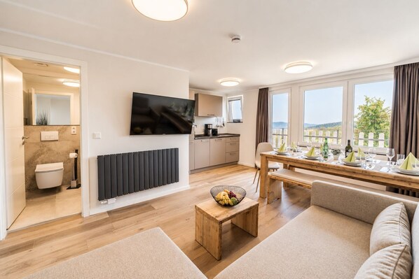 A modern living room with a flat-screen TV, a dining area with a table set for two, and a kitchen in the background.