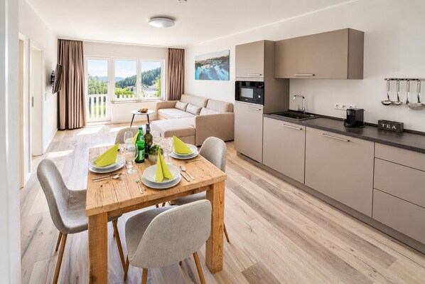 A modern kitchen with a dining area, a sofa, and a view of greenery.