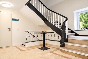 Un escalier avec une rampe noire et des balustrades blanches, menant à un palier où se trouve une petite table et une pancarte au-dessus d’une porte.