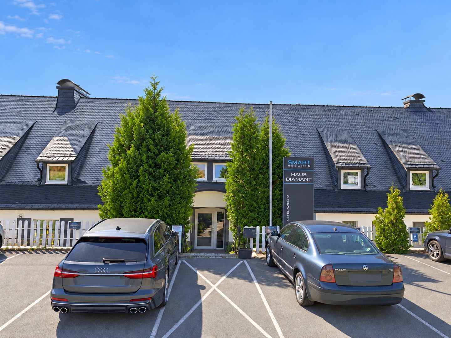 A hotel with a modern design, featuring a parking lot with two cars, a signboard, and a well-maintained lawn.