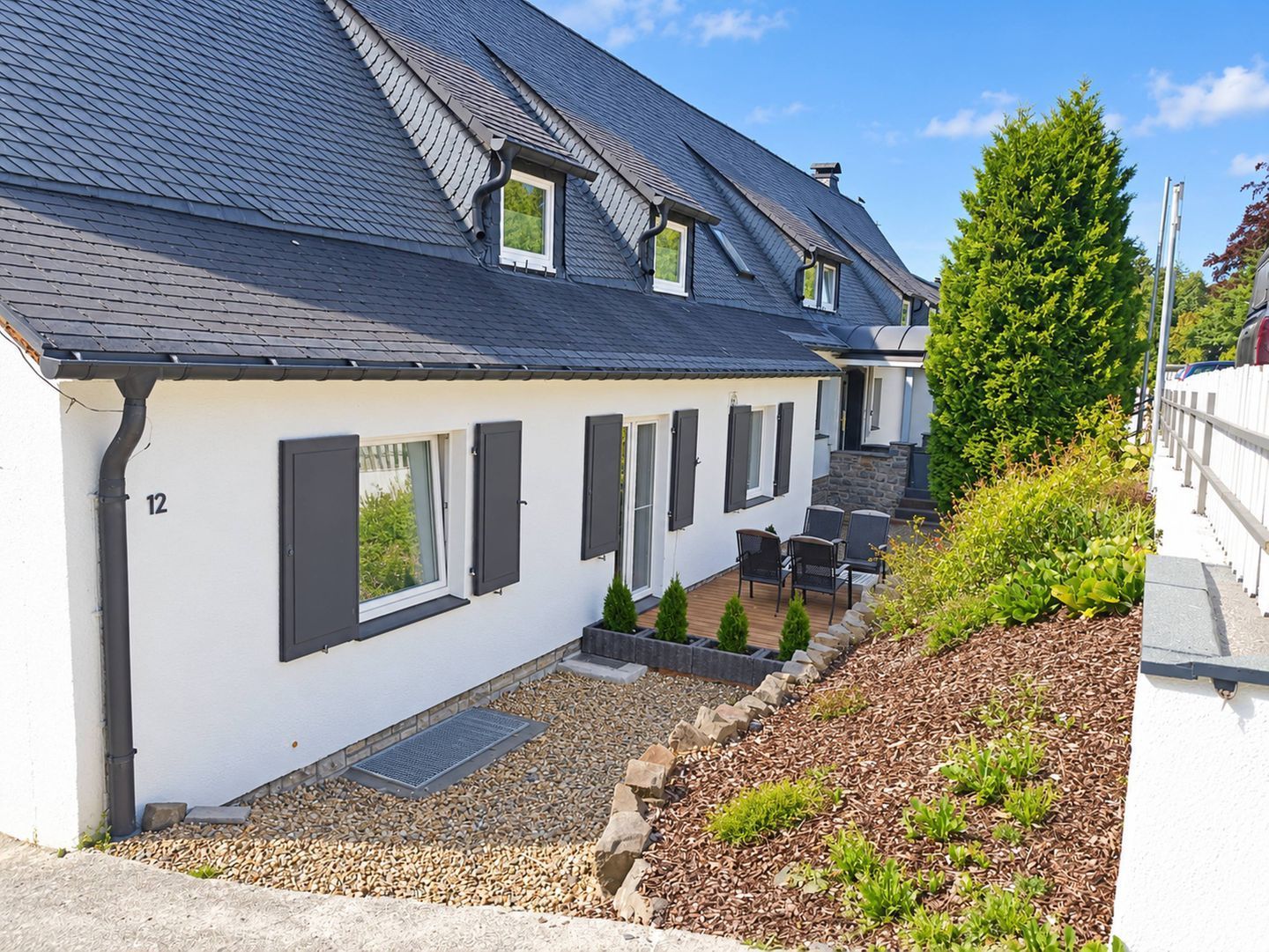 Ein zweistöckiges Haus mit geneigtem Dach, zahlreichen Fenstern mit Fensterläden, einer kleinen Terrasse mit Gartenmöbeln und einem gepflegten Garten.
