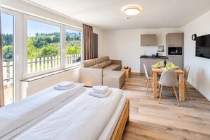 A hotel room with two beds, a sofa, a dining table with chairs, a kitchen area, and a large window with a view of greenery.