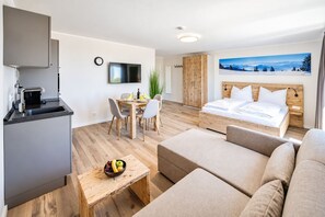 A modern hotel room with a bed, a sofa, a dining table, and a kitchen area.