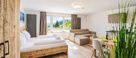 A modern hotel room with a wooden bed, a sofa, a dining table, and a view of the outdoors.