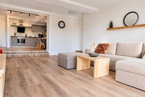 A modern living room with a sofa, ottoman, and coffee table, a kitchen in the background, and a staircase leading to an upper level.