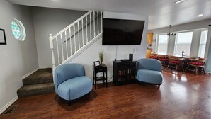 Ein Wohnzimmer mit Treppe, einem an der Wand befestigten Flachbildfernseher und einem Essbereich mit Holztischen und einer Tischplatte.