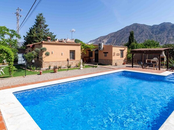 En swimmingpool med en terrasse, en mindre bygning og et bjergrigt landskab i baggrunden.