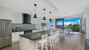 A modern kitchen with a central island, white bar stools, and a view to the outside. - GEORGE8- The Ultimate Family Beach House (Alexandra Headland)