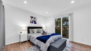 Una camera da letto moderna con un letto grande, un comodino con una lampada, una panca e vista sugli alberi attraverso una porta scorrevole in vetro.