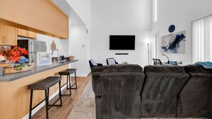 Un soggiorno moderno con un divano grigio scuro, un angolo cottura con mobili in legno, una TV a schermo piatto montata a parete e un quadro astratto decorativo.