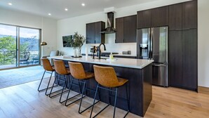 Una cocina moderna con una isla central, taburetes de bar, armarios oscuros, electrodomésticos de acero inoxidable y un amplio ventanal con vistas a zonas verdes.