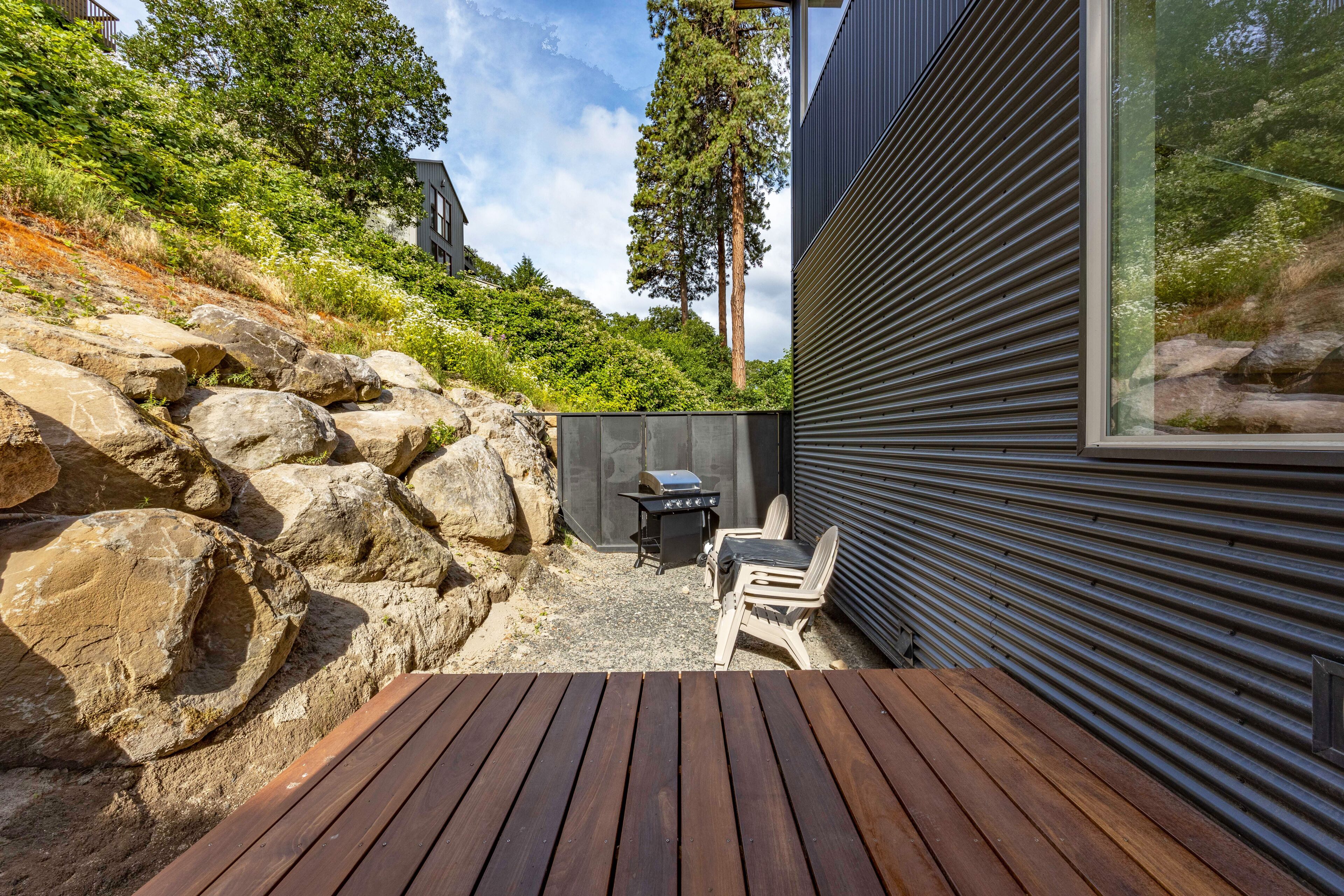 A wooden deck with a grill and chairs, surrounded by large rocks and greenery.