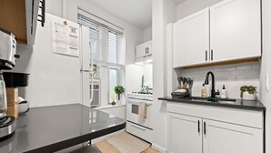 Een moderne keuken met witte kasten, een zwart aanrecht en een fornuis. Er is een raam met jaloezieën, een plant en een welkomstbord aan de muur.
