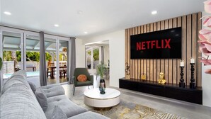 Ein modernes Wohnzimmer mit einem großen Flachbildfernseher an einer Holzwand, einem grauen Ecksofa, einem runden Couchtisch und einer Glastür, die zu einem Außenbereich führt.