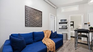 Uma sala de estar moderna com um sofá azul, um painel decorativo na parede e uma área de cozinha com micro-ondas e uma pequena mesa.