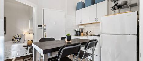 Una cocina compacta con armarios blancos, un comedor con mesa negra y un refrigerador blanco.