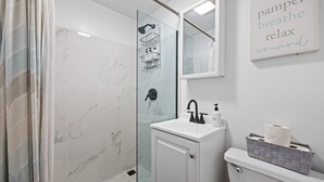 A modern bathroom with a glass-enclosed shower, a white sink with a black faucet, and a toilet with a silver trash bin. - Walk to Time Square! Cozy Studio (New York)