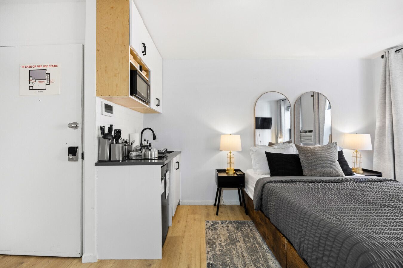 Quarto de hotel moderno com cozinha compacta, cama com colcha cinza, duas luminárias de cabeceira e um espelho grande acima da cama.