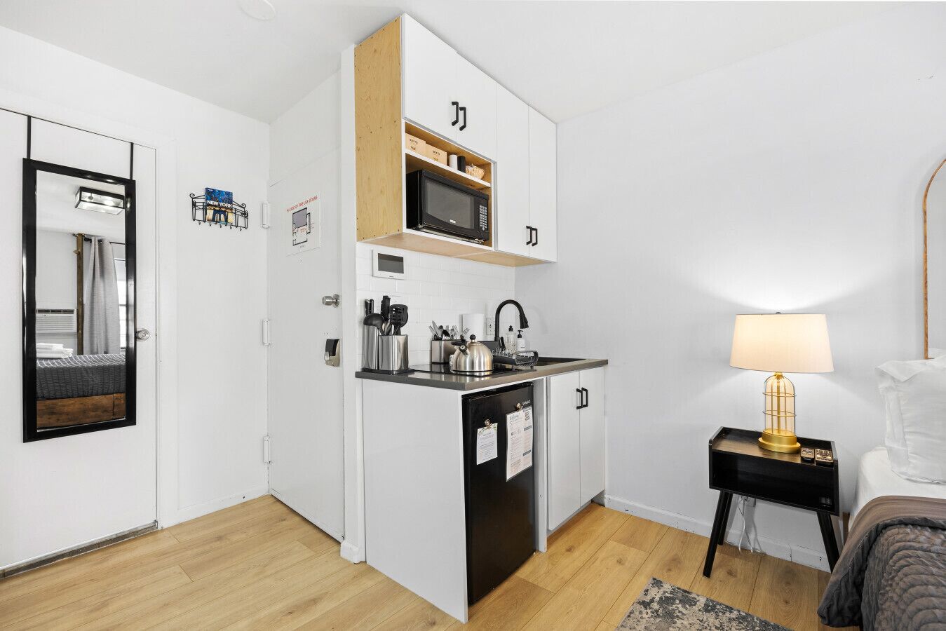Une cuisine compacte avec des meubles blancs, un four à micro-ondes et un évier. On y trouve une petite table avec une lampe, ainsi qu'un lit recouvert d'une couverture grise.