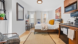 Une chambre d’hôtel de taille réduite, comprenant un lit, une table de chevet, une petite kitchenette, une chaise, un bureau et une fenêtre.