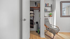 Un couloir mène à une pièce où se trouvent un lit en bois, une chaise, une étagère remplie de livres et une plante en pot.